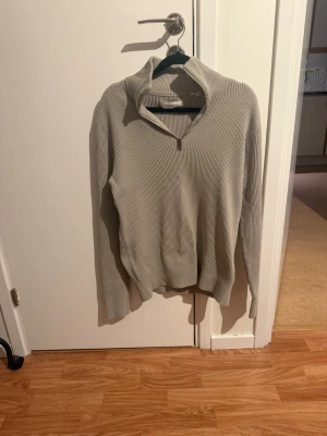 Beige ribbad stickad tröja half zip - Beige stickad tröja med ribbad struktur och half zip-dragkedja i halsen. Tröjan har lång ärm och hög krage, perfekt för lager-på-lager. Enkel och stilren design som passar till många outfits.