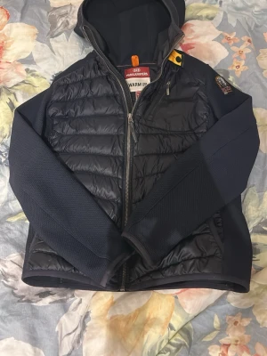 Svart dunjacka från Parajumpers - Snygg svart dunjacka från Parajumpers med huva och dragkedja framtill. Jackan har quiltad kropp, släta ärmar och en gul detalj vid halsen. Märkespatch på ena ärmen och fickor med dragkedja. Perfekt för kalla vinterdagar. Har ett litet hål på ryggen men som är fast strykit med en lite lapp av samma tyg och färg 
