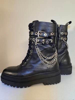 Svarta chunky kängor med kedjor från Zara - Snygga svarta kängor i skinn med grov sula, snörning, spännen och coola silverfärgade kedjor på sidorna. Perfekta för dig som gillar en edgy och alternativ stil. Dragkedja på insidan för enkel på- och avtagning. Inte för breda fötter. 