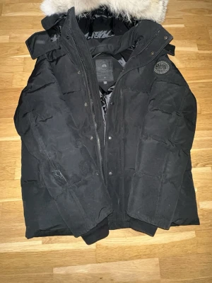 Canada goose wyndham Black Badge - Säljer en svart dunjacka från Canada Goose med avtagbar huva och snygg pälskant. Jackan har patch-logga på ärmen, dragkedja och knappar framtill samt ribbade muddar vid ärmsluten. Perfekt för kalla vinterdagar. Jackan behöver bara rengöras lite sen är den lika bra som ny.