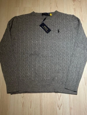 Grå kabelstickad tröja Polo Ralph Lauren - Snygg grå kabelstickad tröja från Polo Ralph Lauren med rund halsringning och klassisk broderad logga på bröstet. Tröjan har ribbade muddar vid ärmslut och nederkant och är tillverkad i mjukt material som känns lyxigt.