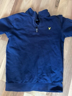 Marinblå halfzip från Lyle & Scott - Snygg marinblå tröja med halv dragkedja från Lyle & Scott. Tröjan har det klassiska gula logobroderiet på bröstet och ribbade muddar. Perfekt för lager-på-lager och enkel att matcha med jeans eller chinos.