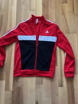 Röd Adidas träningsjacka med dragkedja - Snygg röd träningsjacka från Adidas med vita och svarta detaljer. Jackan har hel dragkedja, ståkrage och klassiska vita ränder längs ärmarna. Tillverkad i ett mjukt och stretchigt material, perfekt för sport eller chill. Adidas-logga på bröstet och Aeroready-tryck bak.