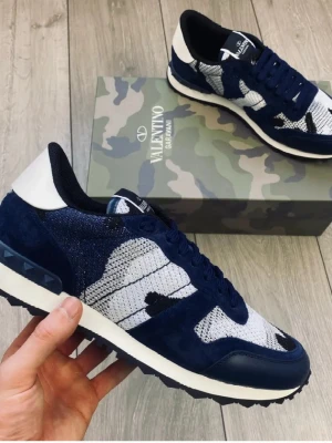 Valentino blå camo sneakers - Snygga sneakers från Valentino med blå och vita camo-detaljer. Ovandelen är i en mix av mocka och textil, med vita och mörkblå partier. Skorna har svarta snören, vit sula och coola nitar på hälen. Perfekta för dig som vill sticka ut med stil.