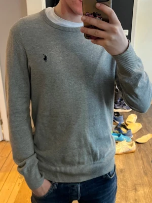 Grå tröja från Ralph Lauren - Snygg grå tröja från Ralph Lauren med klassisk rund halsringning och diskret mörkblå logga på bröstet. Tröjan har ribbade muddar vid ärmslut och nederkant, perfekt för en clean och stilren look. Storleken är Xl ungdom passar S