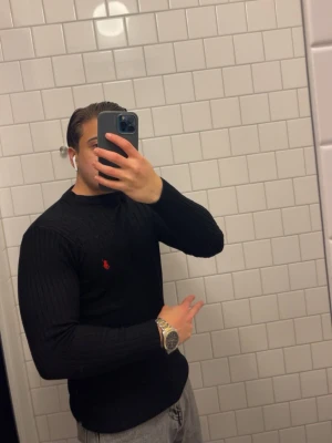 Svart stickad tröja från Ralph Lauren - (Obs dry fit) (Storlek S men den är tight fit så typ Xs) Snygg svart stickad tröja från Ralph Lauren med klassisk röd logga på bröstet. Tröjan har ribbstickad struktur, rund hals och långa ärmar. Perfekt för dig som gillar stilrena och tidlösa plagg.