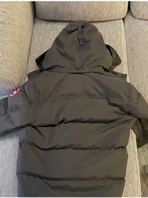 Svart dunjacka från Canada Goose - Säljer en grå dunjacka från Canada Goose med huva och klassisk logga på ärmen. Jackan är vadderad och har en stilren design med raka linjer. Perfekt för kalla dagar när du vill hålla värmen och ändå se snygg ut.
