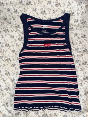 Randig ribbad linnetopp från Levi's - Säljer en ribbad linnetopp från Levi's med marinblå, vita och röda ränder. Toppen är ärmlös och har en liten röd Levi's-logga. STORLEKEN ÄR 14Y!!!