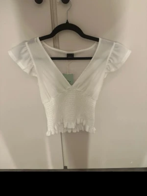 Vit smockad croppad topp från Bik Bok - Superfin vit croppad topp från Gina tricot, med volangdetaljer och v-ringning. Snygg och trendig modell som passar till mycket.🥰👏