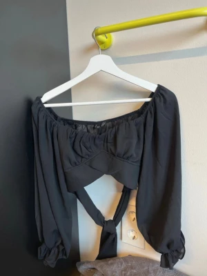 Svart offshoulder topp White Fox - Trendig svart offshoulder topp från White Fox med långa puffärmar och knyt baktill. Tunn och luftig känsla, perfekt för att skapa en cool och chic look. Snygg detalj med resår vid ärmslut och axlar.