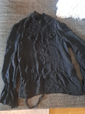 Svart blus med spetsdetaljer - Snygg svart blus med långärmad design och dekorativa spetsdetaljer över bröstet. Blusen har hög hals och knytband i midjan för en feminin siluett. Perfekt att styla med jeans eller kjol för en trendig look.