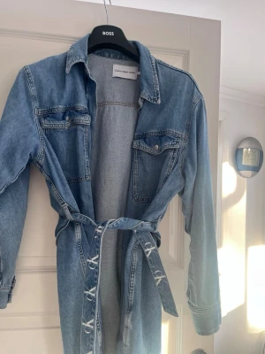 Jeansklänning från Calvin Klein Jeans - Säljer en snygg ljusblå jeansklänning från Calvin Klein Jeans. Klänningen har lång ärm, knappar framtill, två bröstfickor med lock och ett matchande knytskärp i midjan. Perfekt för dig som gillar en avslappnad men trendig stil.