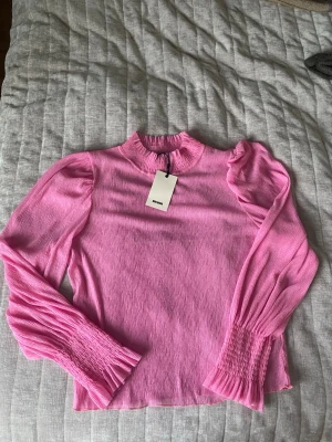 Rosa plisserad blus från Bik Bok - Trendig rosa blus från Bik Bok med plisserat tyg och höga, rynkade muddar vid ärmslut och hals. Blusen har långärmade puffärmar och en lätt glansig finish som ger en cool look. Perfekt att styla med jeans eller kjol för en färgstark outfit. Lite genomskinlig.