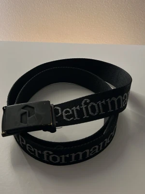 Svart bälte från Peak Performance - Svart bälte från Peak Performance med vit logotyptext längs hela bandet. Bältet har en svart metallspänne med diskret logga. Perfekt accessoar för att ge en sportig touch till din outfit.