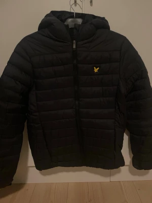 Svart pufferjacka från Lyle & Scott - Snygg svart pufferjacka från Lyle & Scott med gul logga på bröstet. Jackan har huva, dragkedja framtill och quiltad design. Perfekt för kalla dagar och ger en sportig vibe. Materialet är lätt och isolerande.
