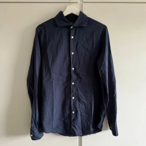 Morris skjorta - Klassisk mörkblå skjorta med vita knappar och lång ärm. Skjortan har en enkel, stilren design och är tillverkad i bomull. Perfekt för dig som gillar en clean och tidlös look.