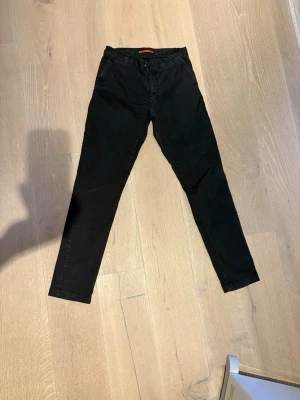 Svarta chinos från Sail Racing - Svarta chinos från Sail Racing med klassisk rak passform och diskreta fickor. Byxorna är tillverkade i ett mjukt bomullstyg som ger en skön känsla och stilren look. Perfekta för dig som gillar en enkel och clean stil. Har även ett par i beige om det är mer intressant! 