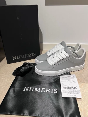 Gråa sneakers från Numeris - Snygga gråa sneakers från Numeris med vita breda skosnören och vit sula. Skorna är alldrig använda, det medföljer även svarta skosnören, skorna är storlek 42 men kan passa 43 denna modell säljs inte längre men når den gjorde gick den för ungefär 2500:-, priset går självklart att diskutera vid snabb affär