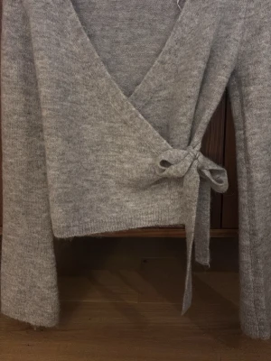 Grå omlottkofta från Gina Tricot - Mysig grå omlottkofta från Gina Tricot i ullblandning. Koftan har långa ärmar, v-ringad hals och knyt i sidan för en snygg passform. Perfekt att slänga på sig när det är lite kyligt ute. Tröjan är använd fåtal gånger.