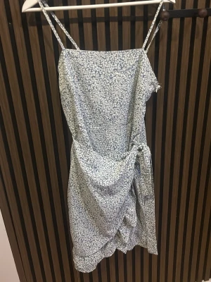 Blommig omlottklänning med smala axelband - Söt kort omlottklänning med blått och vitt blommönster. Klänningen har smala axelband och knyt i sidan för en feminin look. Perfekt för varma dagar och har en lätt, luftig känsla.