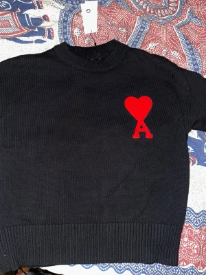 Svart stickad tröja från Ami Paris - Svart stickad tröja från Ami Paris med rund halsringning och ribbade muddar. På bröstet finns ett stort rött hjärta med bokstaven 'A' broderat. Tröjan har en klassisk, rak passform och är tillverkad i ett mjukt stickat material.