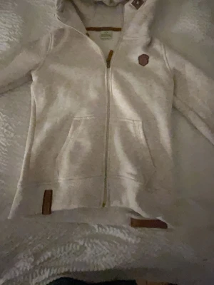 Beige hoodie med dragkedja Naketano - Snygg beige hoodie från Naketano med dragkedja framtill, två fickor och huva. Hoodien har bruna detaljer och märkespatch på bröstet. Mjuk insida och ribbade muddar vid ärmslut och nederkant. Perfekt för en avslappnad och stilren look.