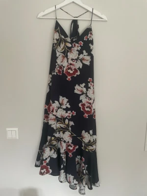 Mörkblå blommig maxiklänning - Säljer en mörkblå klänning med stora blommor i vitt, rött och beige. Klänningen har tunna axelband, knytning i nacken och volangkant nertill. Materialet är tunt och flowy, perfekt för sommardagar. Knappar längs sidan för extra detalj.