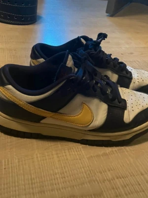 Nike Dunk Low blå/gul/vit sneakers - Säljer ett par Nike Dunk Low sneakers i blått, vitt och gult. Skorna har klassisk låg siluett, perforerad tåbox och gula swoosh-loggor på sidorna. Ovandelen är i läder med mörkblå och vita paneler samt gula detaljer. Perfekta för dig som gillar streetwear.