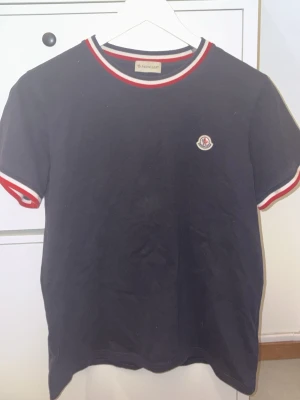 Blå Moncler t-shirt med rödvit kant - Snygg svart t-shirt från Moncler med korta ärmar. T-shirten har rödvita detaljer vid halsringning och ärmslut samt Moncler-logga på bröstet. Tillverkad i mjuk bomull för skön känsla och stilren look.