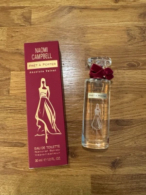 Ny Naomi Campbell Prêt à Porter Absolute Velvet - En elegant parfym från Naomi Campbell, Prêt à Porter Absolute Velvet. Flaskan är genomskinlig med en dekorativ röd rosett vid halsen och har ett stilrent tryck av en kvinna i klänning. Kommer i en vinröd kartong med guldiga detaljer. Innehåller 30 ml 