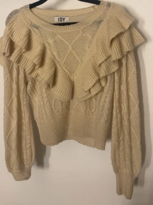 Beige stickad tröja med volanger JDY - Supersöt beige stickad tröja från JDY med volangdetaljer över axlar och bröst. Tröjan har ett kabelstickat mönster, rund halsringning och ribbade muddar vid ärmslut och nederkant. Perfekt för dig som gillar en romantisk och trendig look. knappt använd, inga skador 