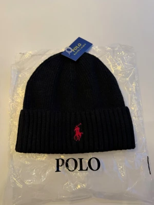 Svart mössa/beanie Polo Ralph Lauren - Snygg svart ribbstickad mössa från Polo Ralph Lauren med uppvikt kant och klassiska röda logobroderiet framtill. Tillverkad i mjukt material som håller dig varm och stilren under kalla dagar. Perfekt accessoar för en trendig streetwear-look. Snabb frakt, skickas samma dag, helt ny samt oanvänd🤗😁