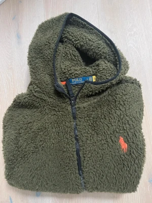 Olivgrön full-zip Hoodie från Polo Ralph Lauren, st. S - Skön olivgrön Curly sherpa full-zip hoodie från Polo Ralph Lauren. Endast använd ett fåtal gånger (för liten för mig som är medium). Jackan har dragkedja framtill, svart kantband och en orange broderad logga på bröstet. Tillverkad i fluffigt teddy-material, perfekt för kalla dagar.