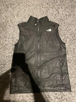 Svart dunväst från The North Face - Snygg svart dunväst från The North Face med hög krage och dragkedja framtill. Västen har två fickor med dragkedja och diskret logga på bröstet och ryggen. Perfekt för lager-på-lager och håller dig varm under kyliga dagar.
