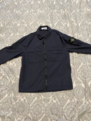 Mörkblå overshirt från Stone Island - Snygg mörkblå overshirt från Stone Island med dragkedja framtill och två bröstfickor. Klassisk krage och ikonisk logotyp-patch på vänster ärm. Tillverkad i ett lätt och slitstarkt material, perfekt för lager-på-lager-stil.