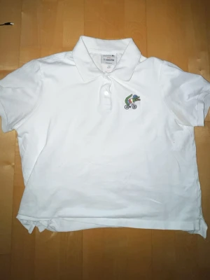 Vit pikétröja Lacoste x Sex Education - Vit pikétröja från Lacoste i samarbete med Netflix Sex Education. Klassisk krage, korta ärmar och liten broderad cyklist på bröstet. Regular fit och tillverkad i mjuk bomull. Perfekt för en clean och sportig look.