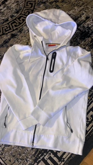 Vit hoodie från Hugo Boss - Snygg vit hoodie från Hugo Boss med dragkedja framtill och svarta detaljer. Hoodien har två fickor med dragkedja och en extra ficka på bröstet. Tillverkad i mjukt bomullsmaterial och har en klassisk huva med dragsko.