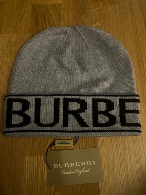 Grå Burberry mössa med logga - Snygg grå mössa från Burberry med bred uppvikt kant och stor svart logotyp runt hela mössan. Tillverkad i mjukt stickat material som håller dig varm och ger en clean streetwear-look. Perfekt accessoar för dig som vill ha något stilrent och trendigt.