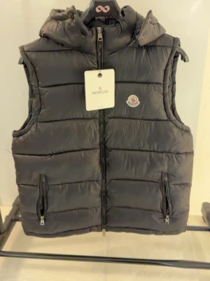 Svart dunväst från Moncler - Snygg svart dunväst från Moncler med huva och två dragkedjeförsedda fickor. Västen har Monclers klassiska logga på bröstet och quiltad design. Perfekt för lager-på-lager och håller dig varm med stil.