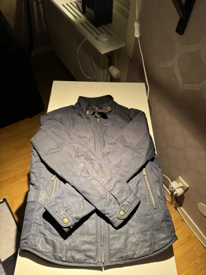 jacka Massimo Dutti - Snygg mörkblå jacka från Massimo Dutti med ståkrage och guldfärgade dragkedjor. Jackan har två framfickor med dragkedja och detaljer i läder vid kragen. Tillverkad i polyester och har en quiltad look. Stlk M. Perfekt som en vårjacka.