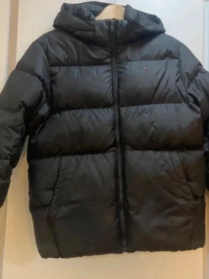 Svart pufferjacka från Tommy Hilfiger - Säljer en svart pufferjacka från Tommy Hilfiger med huva och dragkedja framtill. Jackan har en diskret logga på bröstet och breda, vadderade paneler som ger en riktigt fet look. Perfekt för kalla dagar när du vill hålla värmen och ändå se stilren ut.