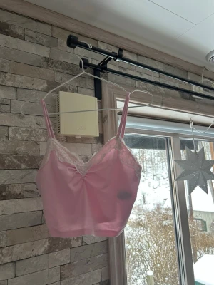 Rosa linne  med smala axelband - Söt rosa linne  med smala axelband och vit spetsdetalj längs urringningen. Mjuk och stretchig modell som sitter bekvämt mot kroppen. Perfekt för dig som gillar en feminin och enkel stil.