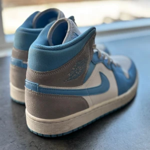 Nike Air Jordan 1 Mid blå/vit/grå - Snygga Nike Air Jordan 1 Mid sneakers i blått, vitt och grått. Skorna har klassisk hög siluett, ikonisk swoosh på sidan och vingar-logga. Ovandelen är i mocka och läder för en clean och stilren look. Perfekta för dig som vill sticka ut med streetwear-vibe.