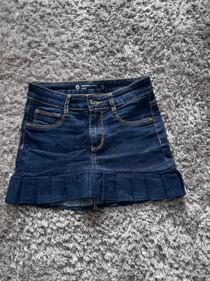 Mörkblå jeanskjol med volangkant - Säljer en mörkblå jeanskjol från Jordan Jean med volangkant nedtill och söta rosettbroderier på bakfickorna. Kjolen har klassiska fickor fram och bak samt kontrastsömmar. Perfekt för dig som gillar en söt och trendig stil. Den är i s men skulle säga att den är mer en xs eller liten s 