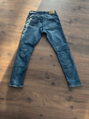 Blå slim fit jeans med slitningar - 33/32 Säljer ett par blå slim fit jeans med snygga slitningar och tvättade detaljer. Jeansen har fem fickor, normal midja och klassisk knappgylf. Passar perfekt till en avslappnad streetwear-look.