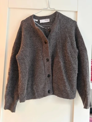 Grå stickad kofta från Selected Femme - Mysig grå stickad kofta från Selected Femme med rund halsringning och knäppning framtill med fem svarta knappar. Koftan har lång ärm och är tillverkad i ett mjukt, ulligt material som känns riktigt skönt mot huden. Perfekt för lager på lager och enkel att styla.