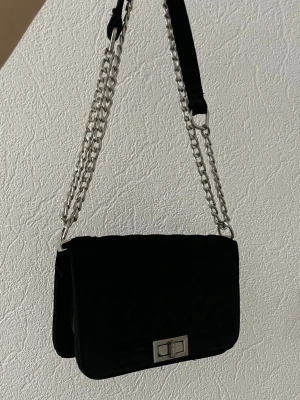 Svart quiltad axelväska med kedja - Snygg svart axelväska i quiltat tyg med silverfärgad kedjerem och coolt vridlås framtill.                                MAN KAN ÄVEN FÖRLÄNGA DEN eller ha som en vanlig axelväska (kortare). Lite smutsigt inne men går att torka, tvätta bort det. Pris kan diskuteras 💞