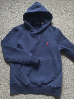 Blå hoodie från Polo Ralph Lauren - Mörkblå hoodie från Polo Ralph Lauren i slim fit-modell. Klassisk design med huva, magficka och broderad röd logga på bröstet. Tillverkad i mjuk bomullsblandning som är perfekt för en avslappnad och stilren look. Storlek S men passar även xs