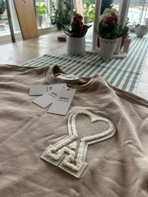 Beige sweatshirt från Ami Paris - Snygg beige sweatshirt från Ami Paris med stort vitt hjärta och A-logga broderat på bröstet. Tröjan har rund halsringning, ribbade muddar och är tillverkad i mjuk bomull. Perfekt för en avslappnad och trendig look.