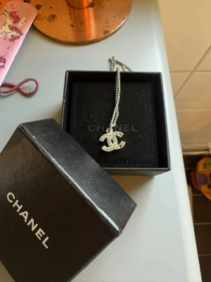Chanel halsband med pärldetaljer - Snyggt halsband från Chanel med den ikoniska CC-loggan som hänge. Hänget är dekorerat med små pärlliknande stenar och kedjan är i silverfärg. Perfekt accessoar för att lyfta din outfit med en lyxig touch. OBS, halsbandet saknar två stenar (därav priset!) 